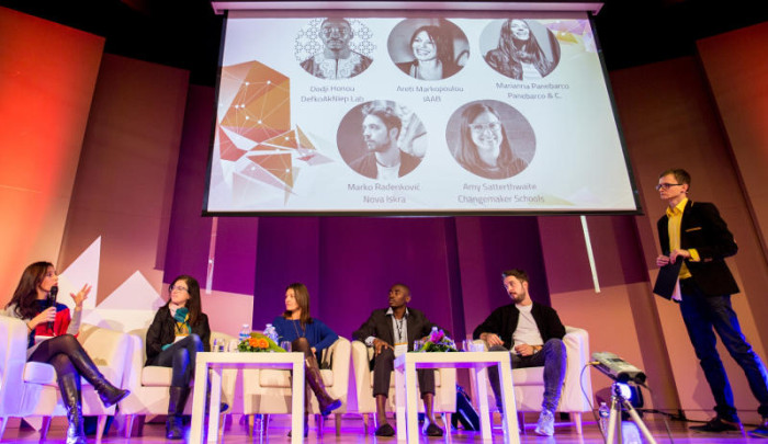 Photograph showing five seated contributors and one standing moderator on stage at a b.creative event.