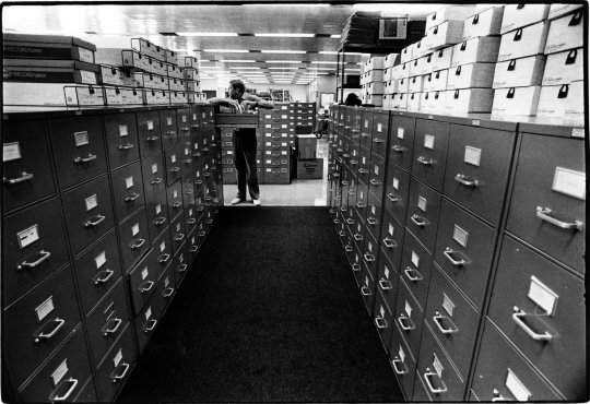 A corridor with filing cabinets and archive boxes lined and stacked on either side.