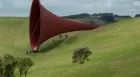 Anish Kapoor structure of a giant ear horn or speaker, laid on a green hillside. Tiny people surround the mouth of it, as though they could walk inside.