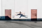 An acrobat in a white tank top and blue jeans performs a straddle jump.