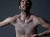 A white man with one arm bent, one arm stretched looks strongly into the camera against a grey, dusky background.