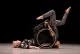 Two women, one in a wheelchair that's tipped over so she's flat on her back. The other holding to the frame of the chair as she stands on her head.