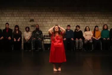 Photograph from Saša Asentic's Dis Sylphide. A woman in a red dress has her hands at her head, running through her hair. In the background, in soft focus, a line of audience seated against a wall.