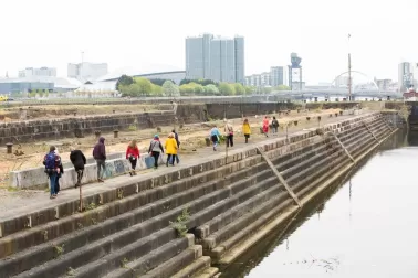 Photograph showing a line of people on a pier, walking towards a city in the distance - Glasgow. Grey sky.