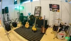 Three gleaming guitars rest on stands in a small music studio which is also filled with speakers, keyboards, a drum kit, and other equipment.