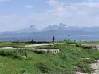 A man stands on a stone platform in a grassy field by the sea. Mountains are distantly visible on an opposite shore.