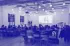 An audience of around thirty sit watching a lecture given over Zoom and projected on a wall.