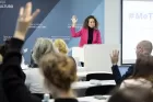 At a lectern in a conference hall a woman in front of a mic is standing with her hand raised. A few members of the audience also have raised hands.