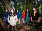 Photo of 6 people in the woods, 5 men in plain clothing and a woman in plain clothing in the foreground with a flower crown