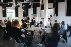 Classic seminar photo: lots of people seated round small tables, man and woman at front beside a projection screen and a flip chart.