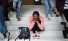 A young woman sitting on a set of concrete stairs, wearing a face mask, looking directly to camera. People are streaming past her, but we only see their legs.